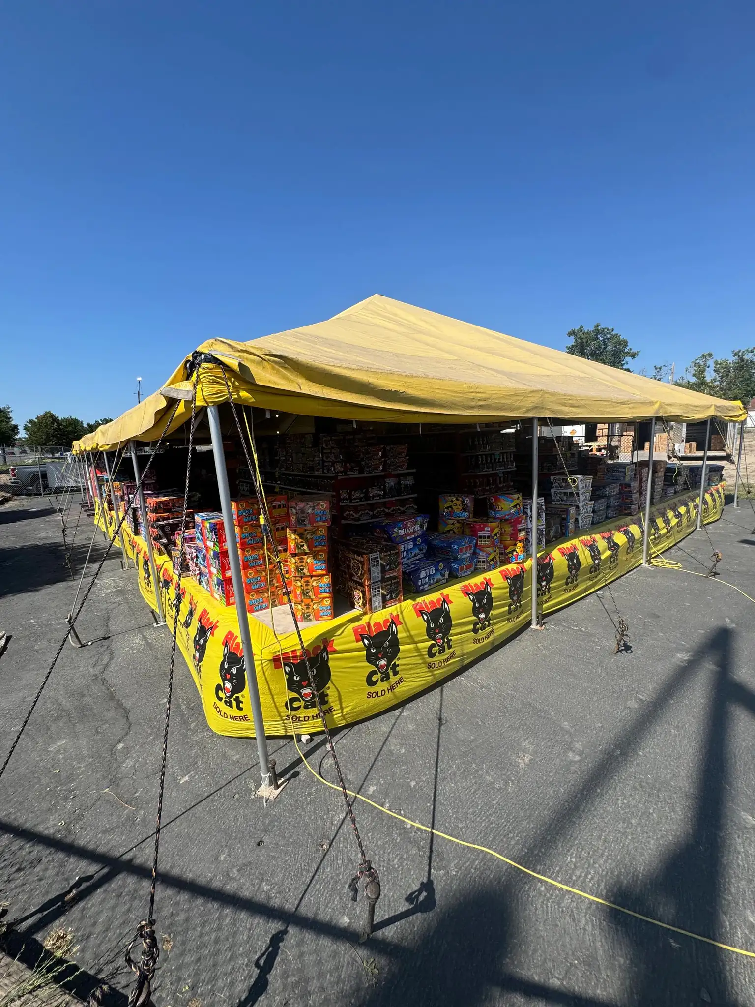 A yellow fireworks tent on a lot, stock stacked under the canopy against a clear blue sky